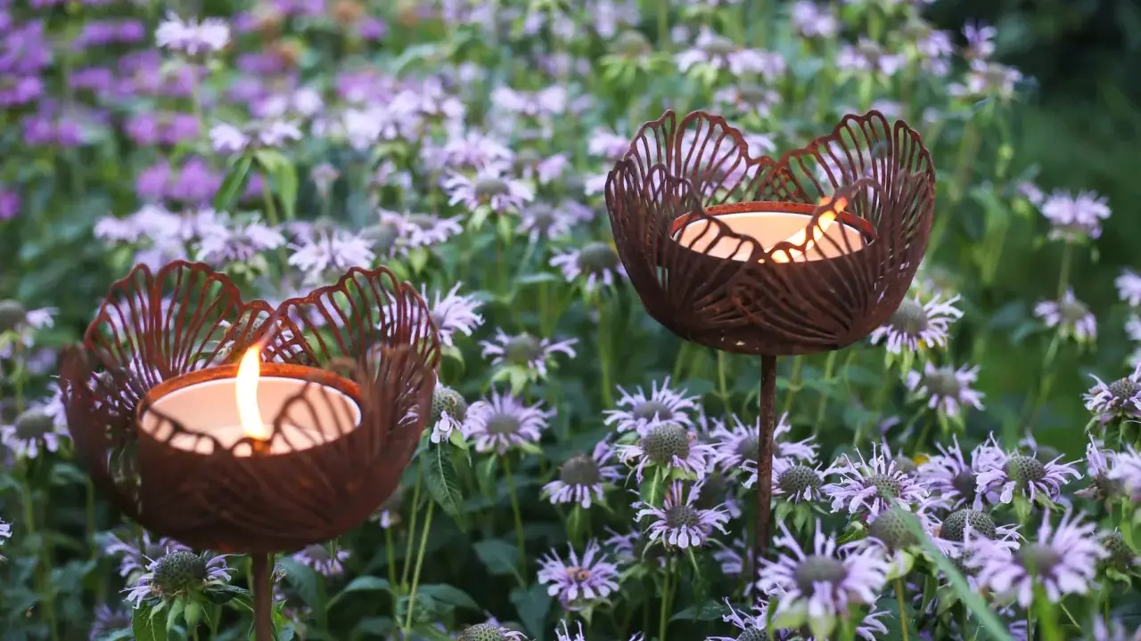 Två rostiga ljushållare i form av blommor lyser upp en rabatt med lila blommor.