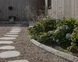En stenlagd gång leder genom grus och flis till gångar, kantad av frodiga hortensior och prydnadsgräs mot en träfasad.