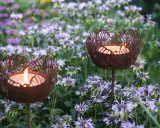 Två rostiga ljushållare i form av blommor lyser upp en rabatt med lila blommor.