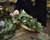 Händer som pysslar med att binda julkrans av granris och gröna blad.