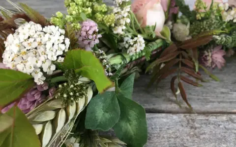 Vacker krans med vita och rosa blommor, gröna blad och ax. Någon har valt att binda krans med kärlek.