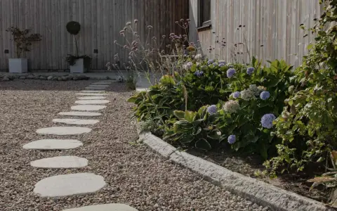 En stenlagd gång leder genom grus och flis till gångar, kantad av frodiga hortensior och prydnadsgräs mot en träfasad.