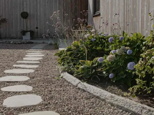 En stenlagd gång leder genom grus och flis till gångar, kantad av frodiga hortensior och prydnadsgräs mot en träfasad.