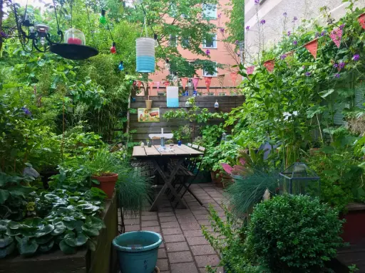 En lummig innergård med ett träbord och stolar, omgivet av grönska. En **klematis i kruka** klättrar längs staketet, och färgglada lyktor hänger från träden.