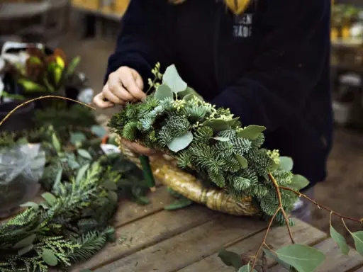 Händer som pysslar med att binda julkrans av granris och gröna blad.