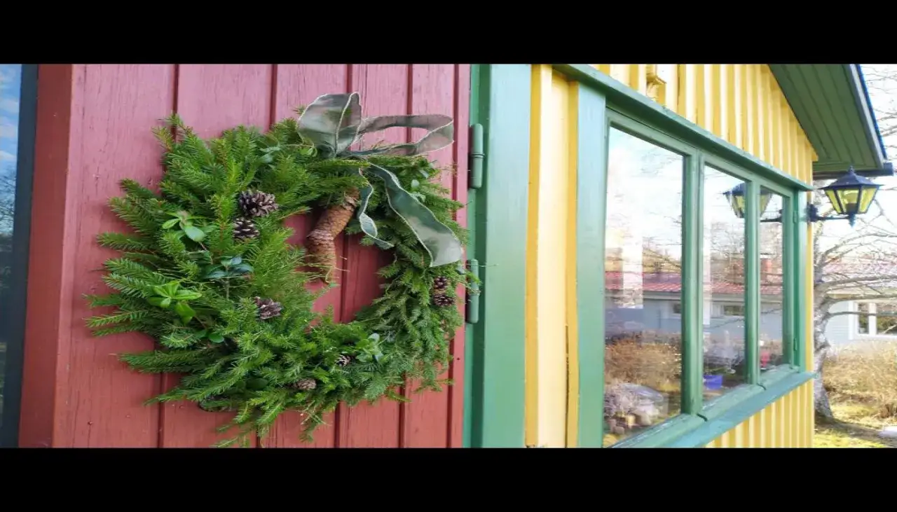 Vacker julkrans med kottar och gr&ouml;nt band, redo att binda julst&auml;mning p&aring; en r&ouml;d d&ouml;rr.