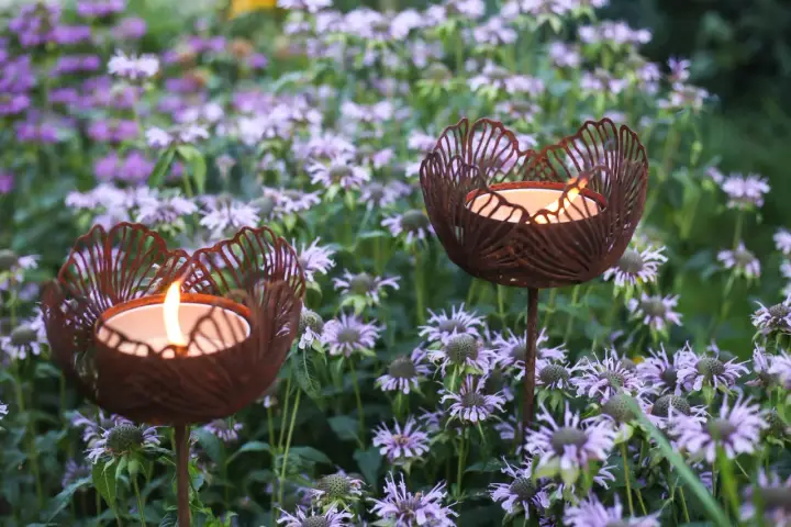 Två rostiga ljushållare i form av blommor lyser upp en rabatt med lila blommor.